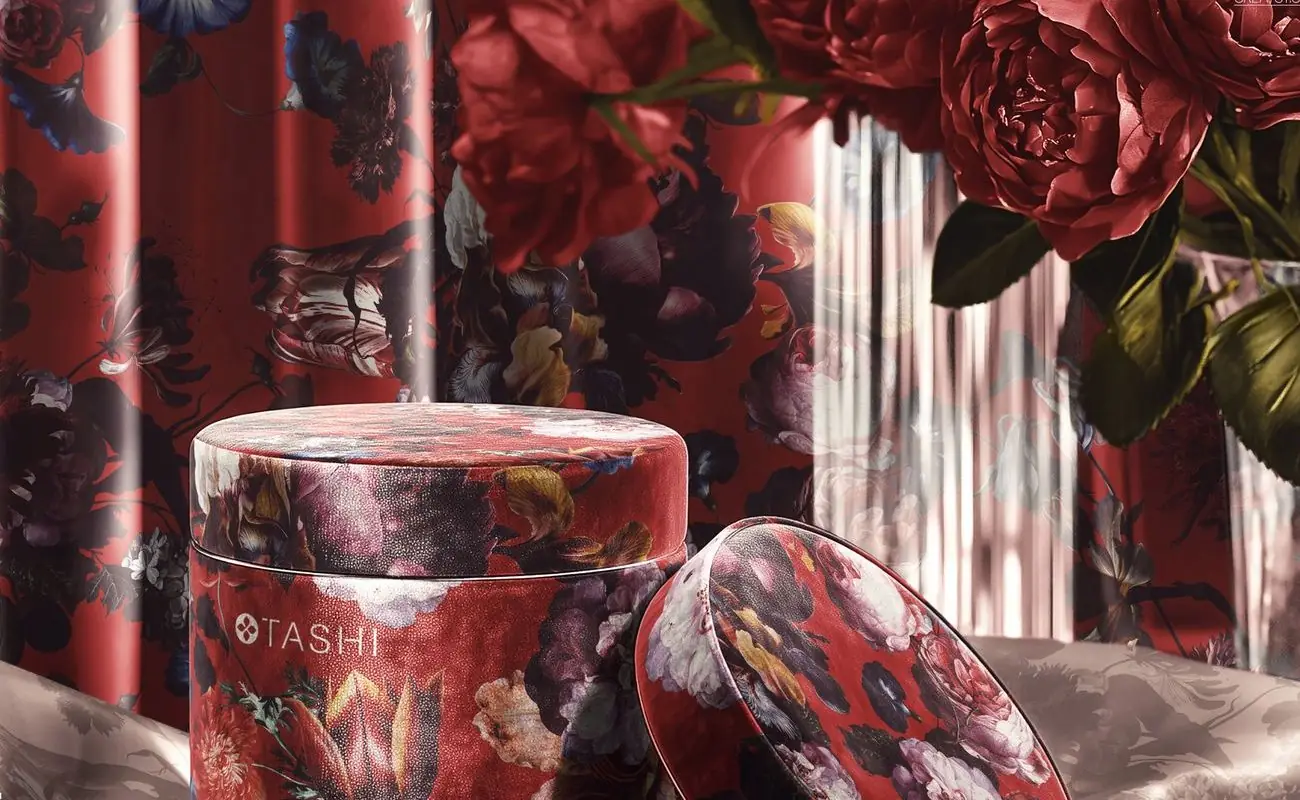 A round velvet-finish gift box printed with a dark floral pattern, styled with deep-red roses and draped satin.